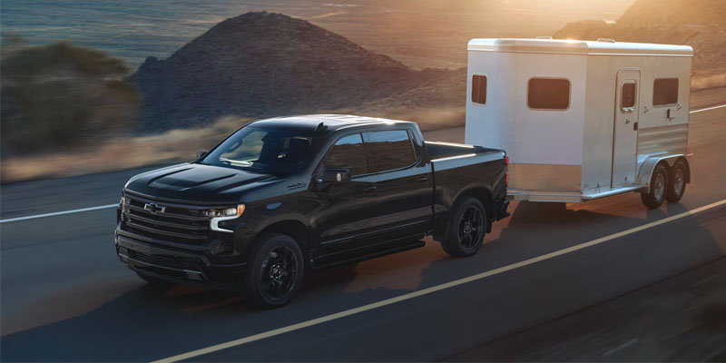 A black Chevrolet Silverado 1500 towing a silver trailer on a highway near rocky hills during sunset.