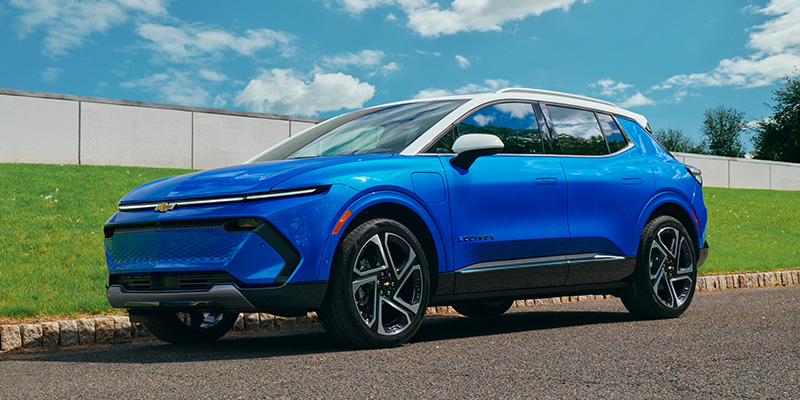 A blue Chevrolet electric SUV parked on a road beside grassy terrain and a partly cloudy sky.